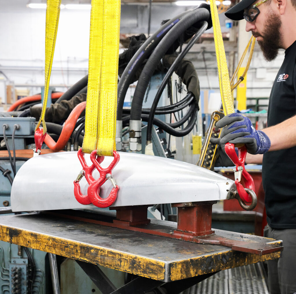 Factory worker wearing safety gear secures a large metal part with red hooks and yellow straps onto a hydraulic lift table in an industrial setting.